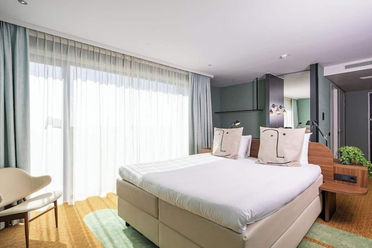Una camera d'albergo moderna con un letto grande, una scrivania, una sedia e un'ampia finestra con tende.