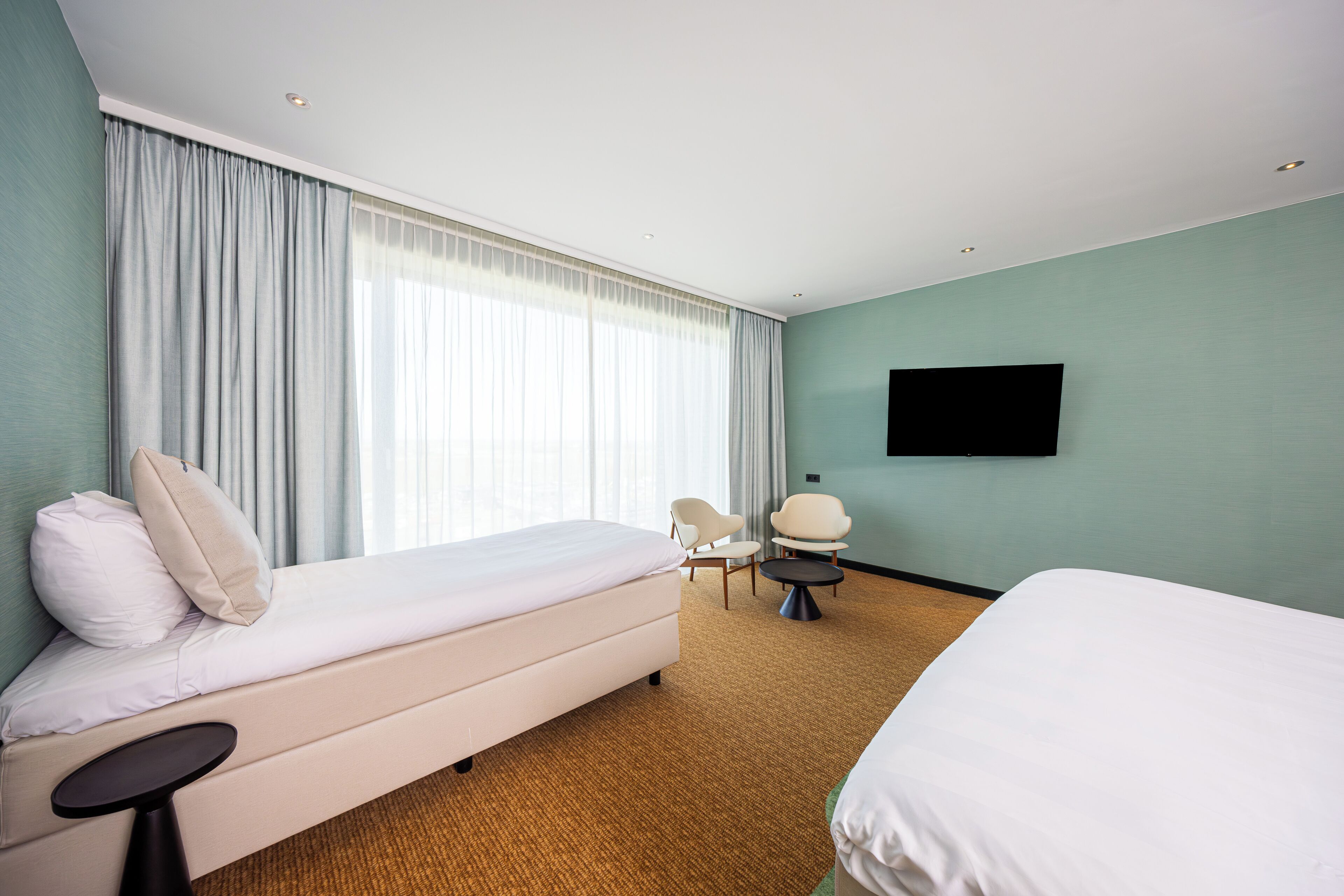 Una habitación de hotel con cama, una silla, una mesita, televisor y un ventanal con cortinas.