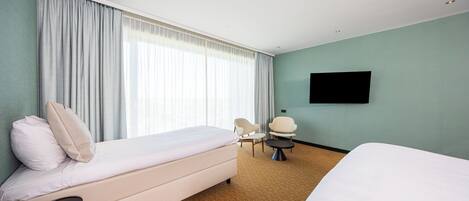 Une chambre d’hôtel avec un lit, une chaise, une petite table, un téléviseur et une grande fenêtre avec des rideaux.