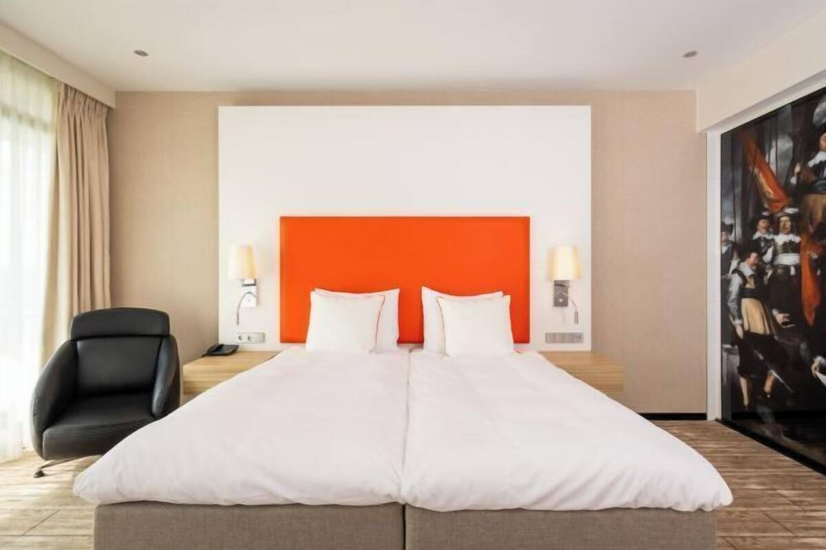 Una camera d'albergo con un letto grande, una sedia, un quadro al muro e una finestra con tende.