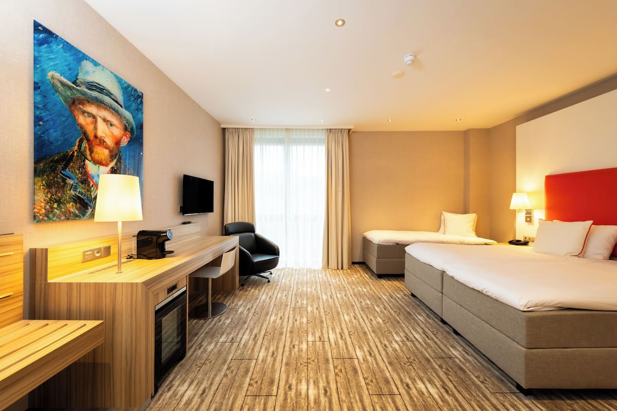 Camera d'albergo con pavimento in legno, un letto, una scrivania con lampada, una sedia, una TV e un quadro appeso al muro.
