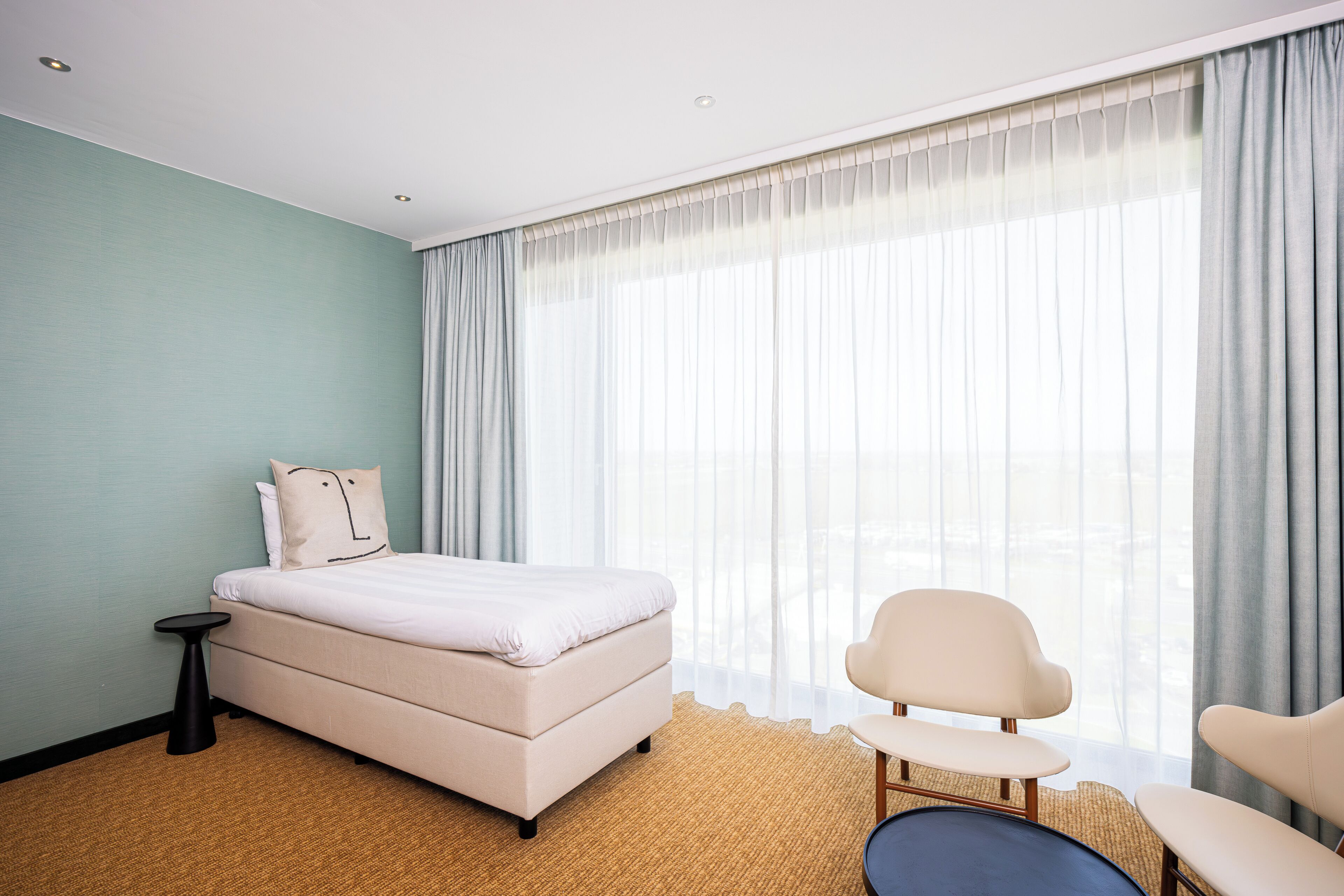 Una habitación de hotel moderna con una cama, una mesita auxiliar, una silla y un ventanal con cortinas.