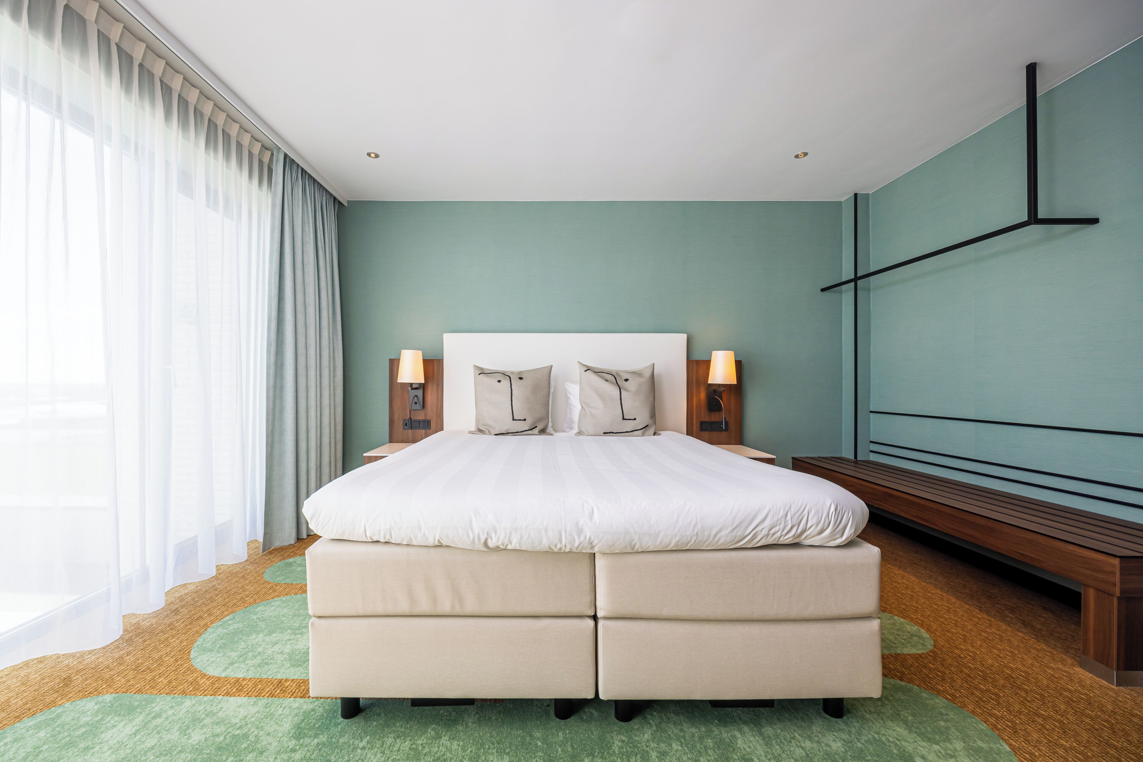 Habitación de hotel moderna con una cama grande, dos lámparas de noche, un banco de madera y una alfombra verde.