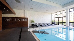 A modern indoor swimming pool with a 'WELLNESS' sign, sun loungers, and a view of the outside.