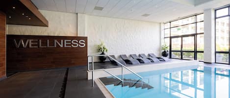 A modern indoor swimming pool with a 'WELLNESS' sign, sun loungers, and a view of the outside.