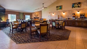 Un comedor de hotel con varias mesas y sillas, una zona de cocina con una cafetera, y un amplio ventanal que proporciona luz natural.