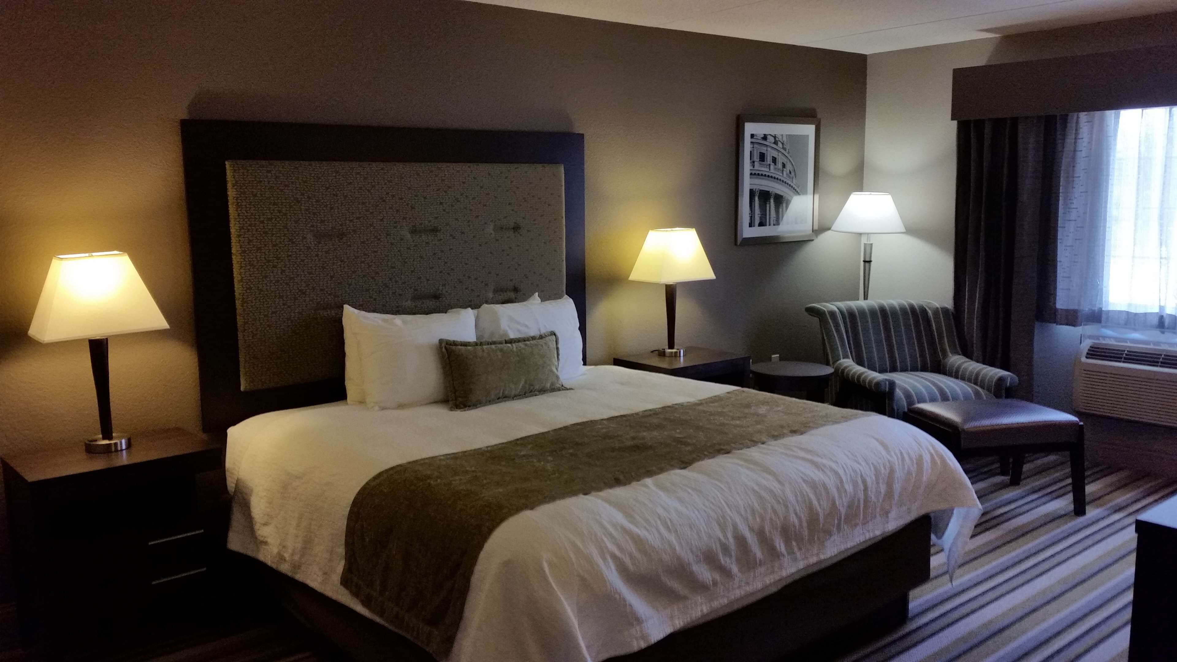 Habitación de hotel con una cama grande, dos lámparas de noche, una silla, una mesita y un cuadro enmarcado en la pared.