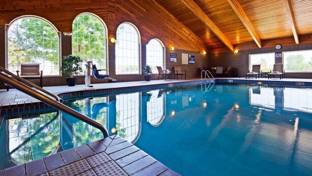 Una piscina interior con paredes de vidrio transparente, vigas de madera en el techo y una zona de estar junto a la piscina con sillas y plantas en macetas.