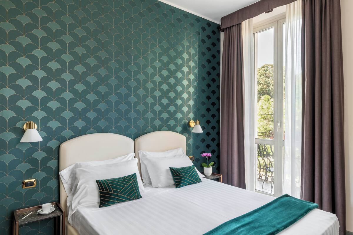 Una camera d'albergo con un letto, due comodini, una finestra con tende e una parete decorata.