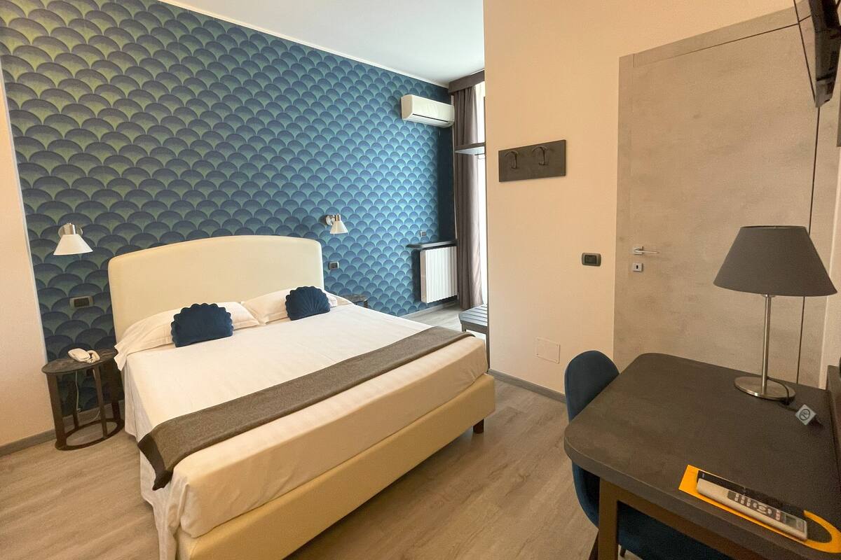 Una camera d'albergo con un letto, una lampada da comodino, una scrivania, una sedia e una parete con motivo geometrico.