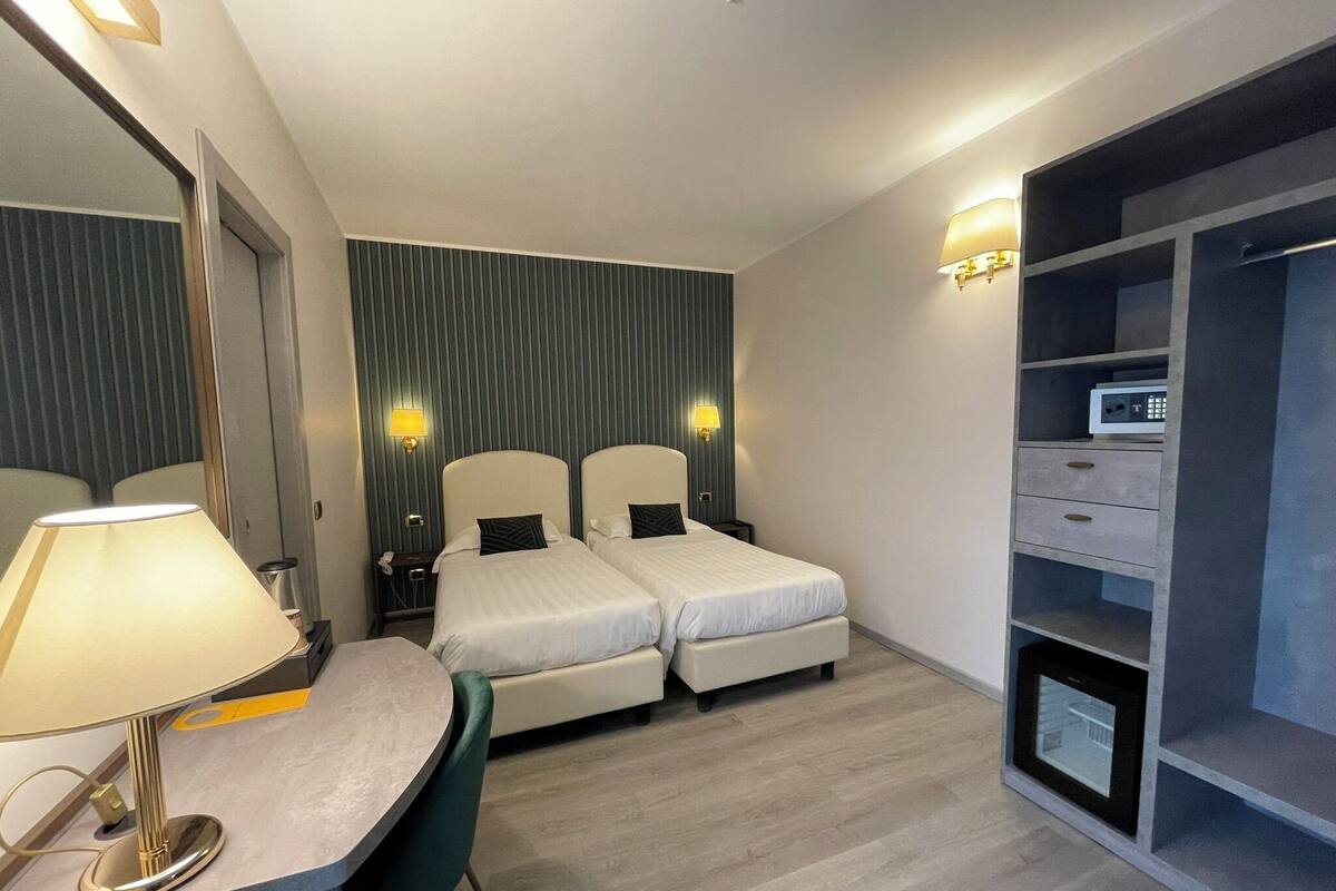 Una camera d'albergo con due letti, una scrivania con lampada, un armadio e uno specchio.