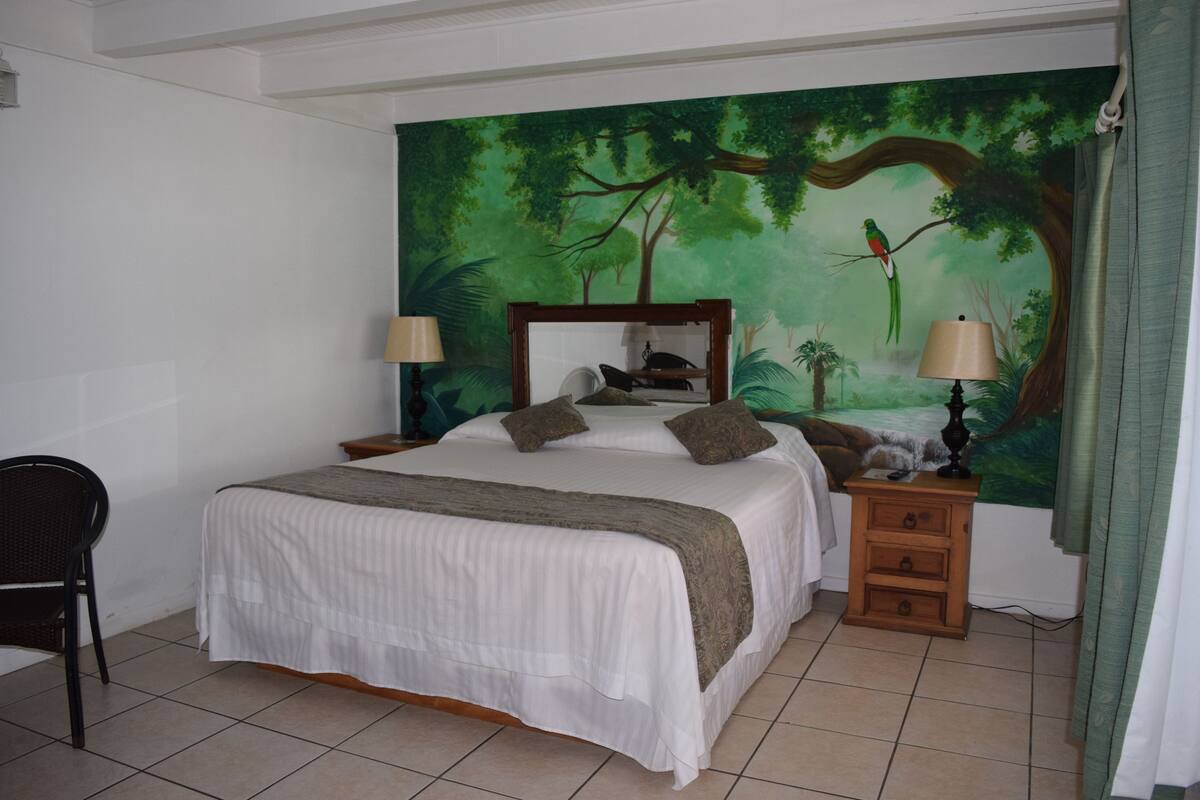 A bedroom with a bed, two nightstands, a chair, and a large forest-themed mural on the wall.