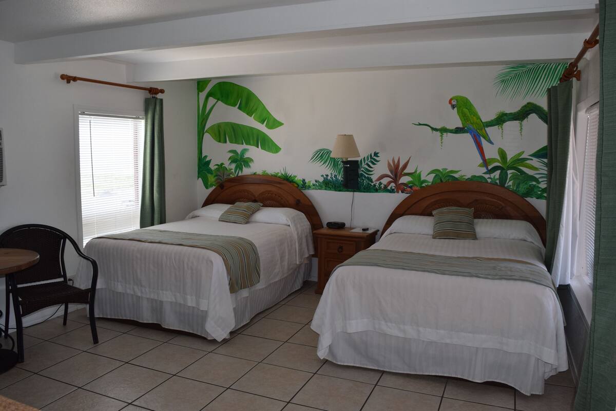 A room with two beds, a chair, a table, and a mural of tropical plants and a parrot.