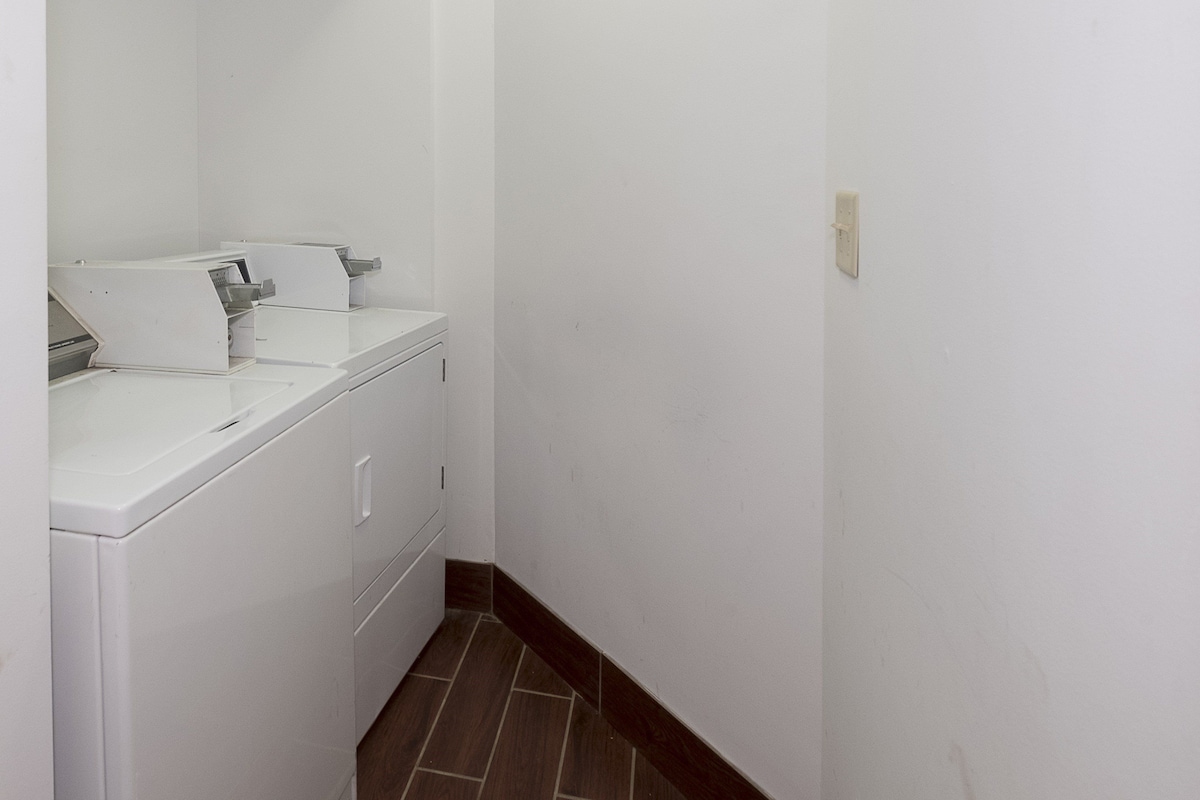A narrow hallway with a washing machine and a dryer on the left, white walls, and a brown tiled floor.
