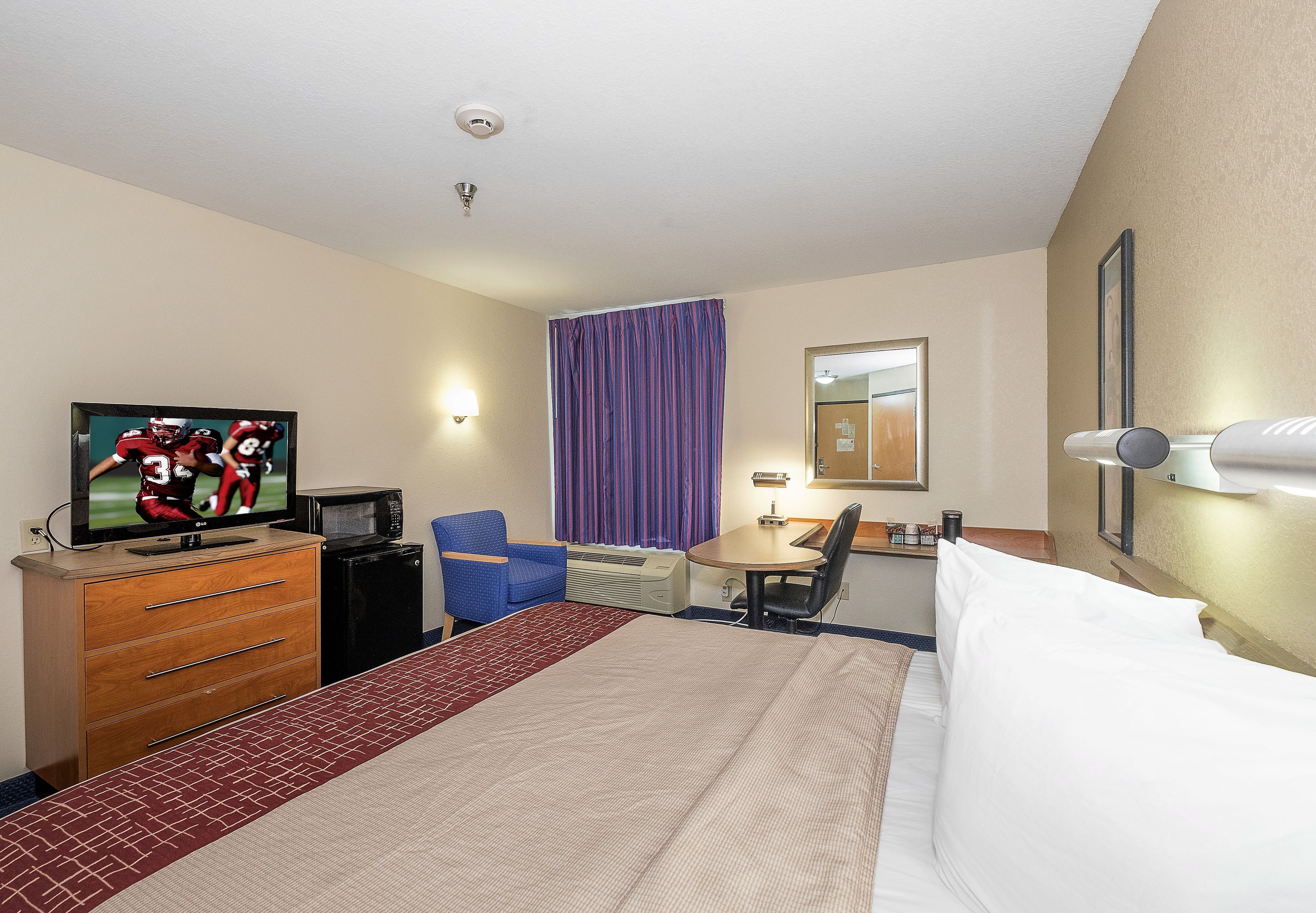Een hotelkamer met een bed, bureau, stoel, televisie, magnetron en raam met gordijnen.