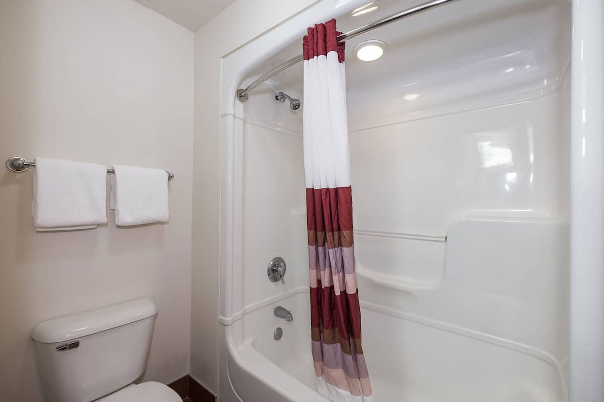 A bathroom with a white toilet, two white towels on a rack, a shower with a red and white curtain, and a white bathtub.