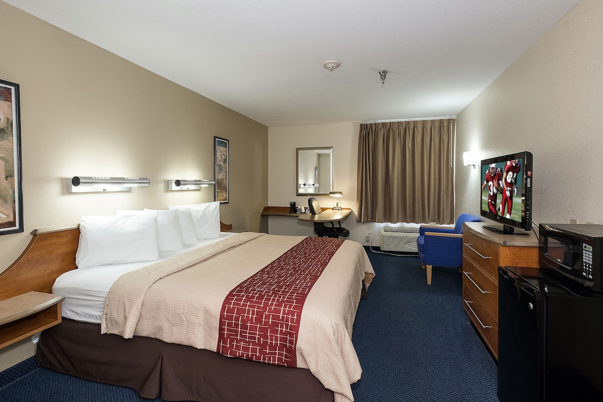 A hotel room with a bed, a desk with a television, a chair, a desk lamp, and a window with curtains.