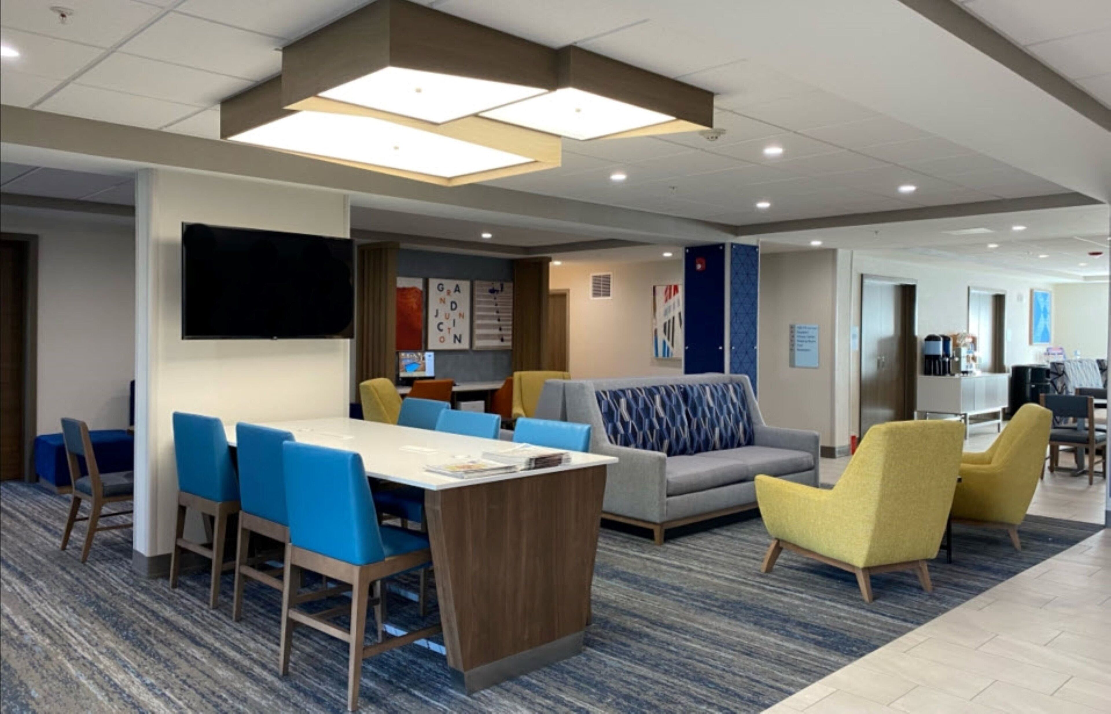 Een moderne hotellobby met een grote tafel, blauwe en gele stoelen, een bank en kunstwerken aan de muren.