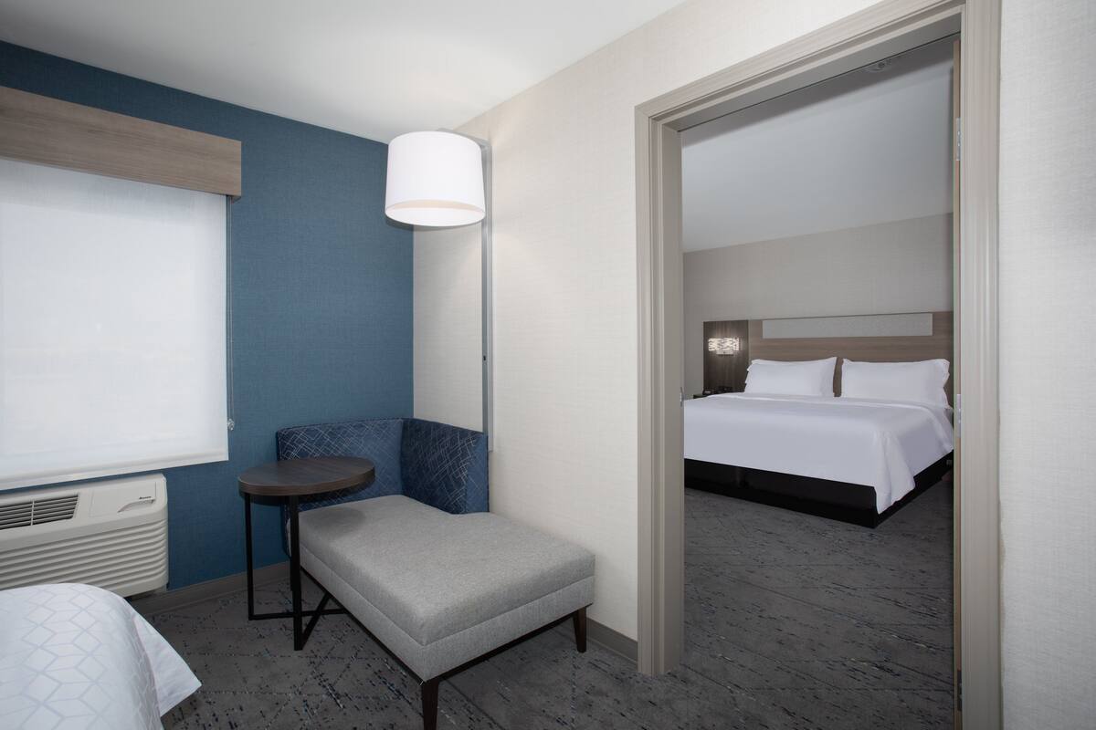 A hotel room with a bed, a seating area with a chair and a small table, a standing lamp, and a window with blinds.