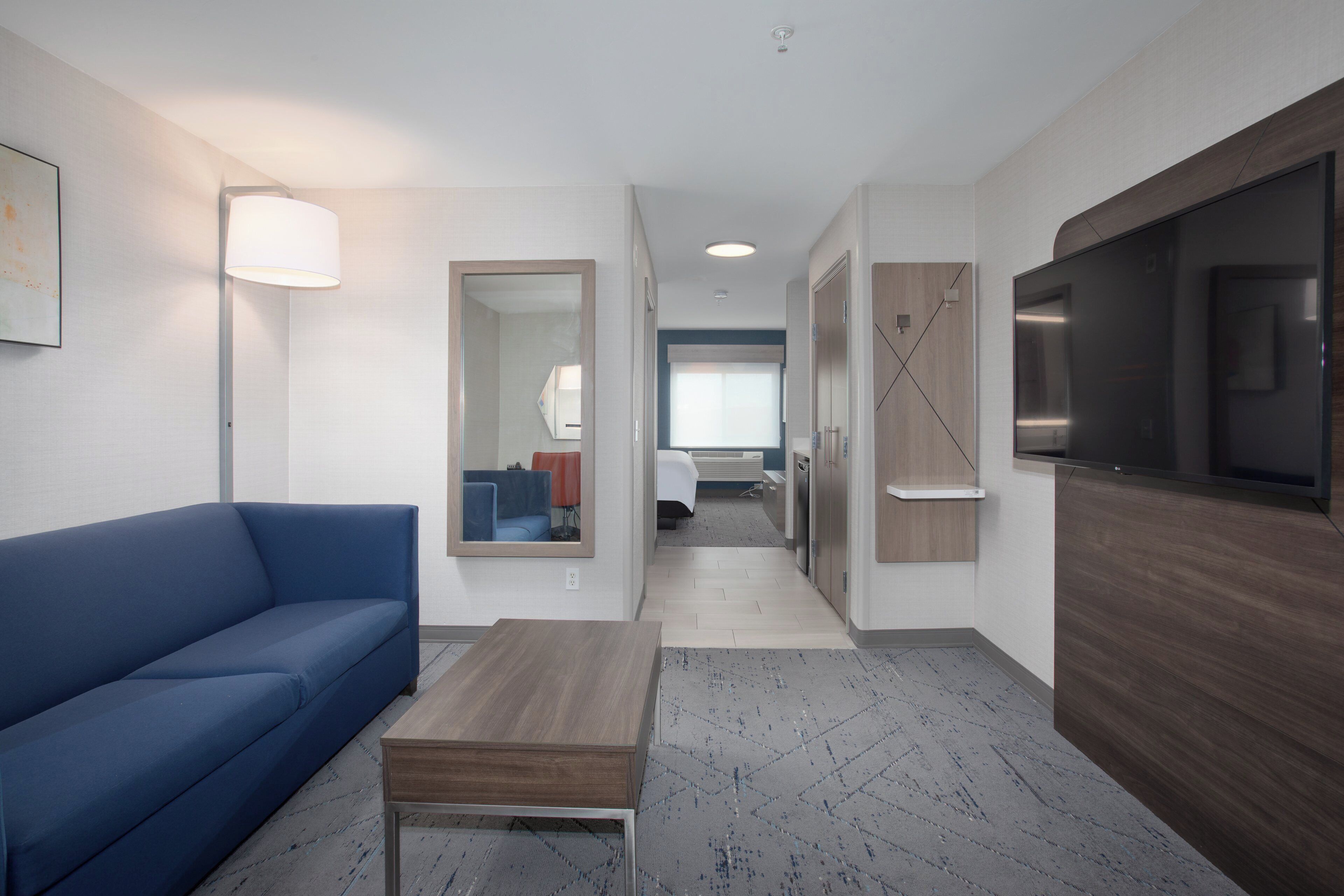 A hotel room with a blue sofa, a wooden coffee table, a flat-screen TV, a mirror, and a hallway leading to a bathroom.