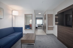 Ein Hotelzimmer mit einem blauen Sofa, einem Holztisch, einem Flachbildfernseher, einem Spiegel und einem Flur, der zu einem Badezimmer führt.