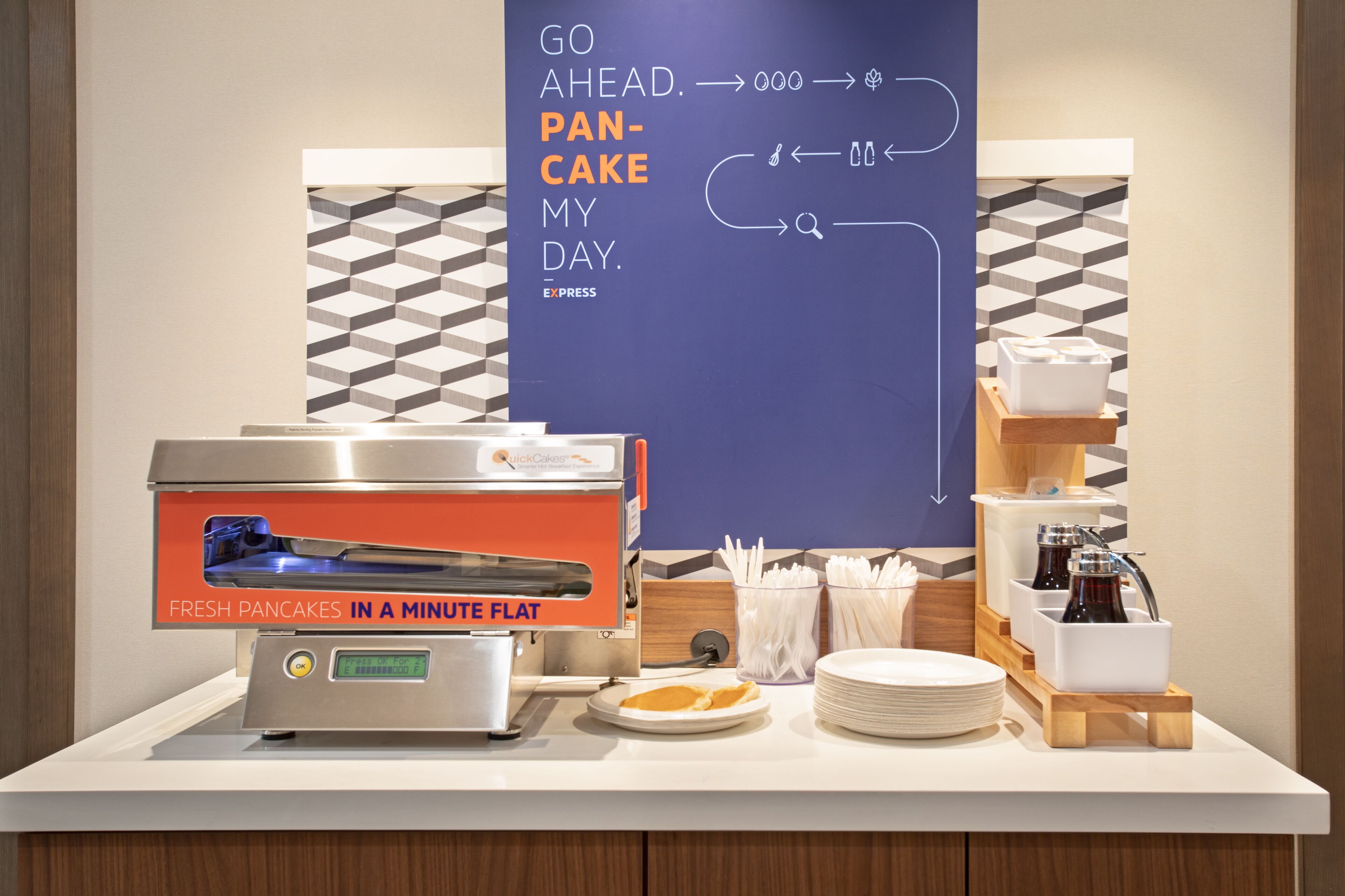 A breakfast station with a pancake maker, plates, napkins, and a sign encouraging guests to "GO AHEAD. PANCAKE MY DAY."