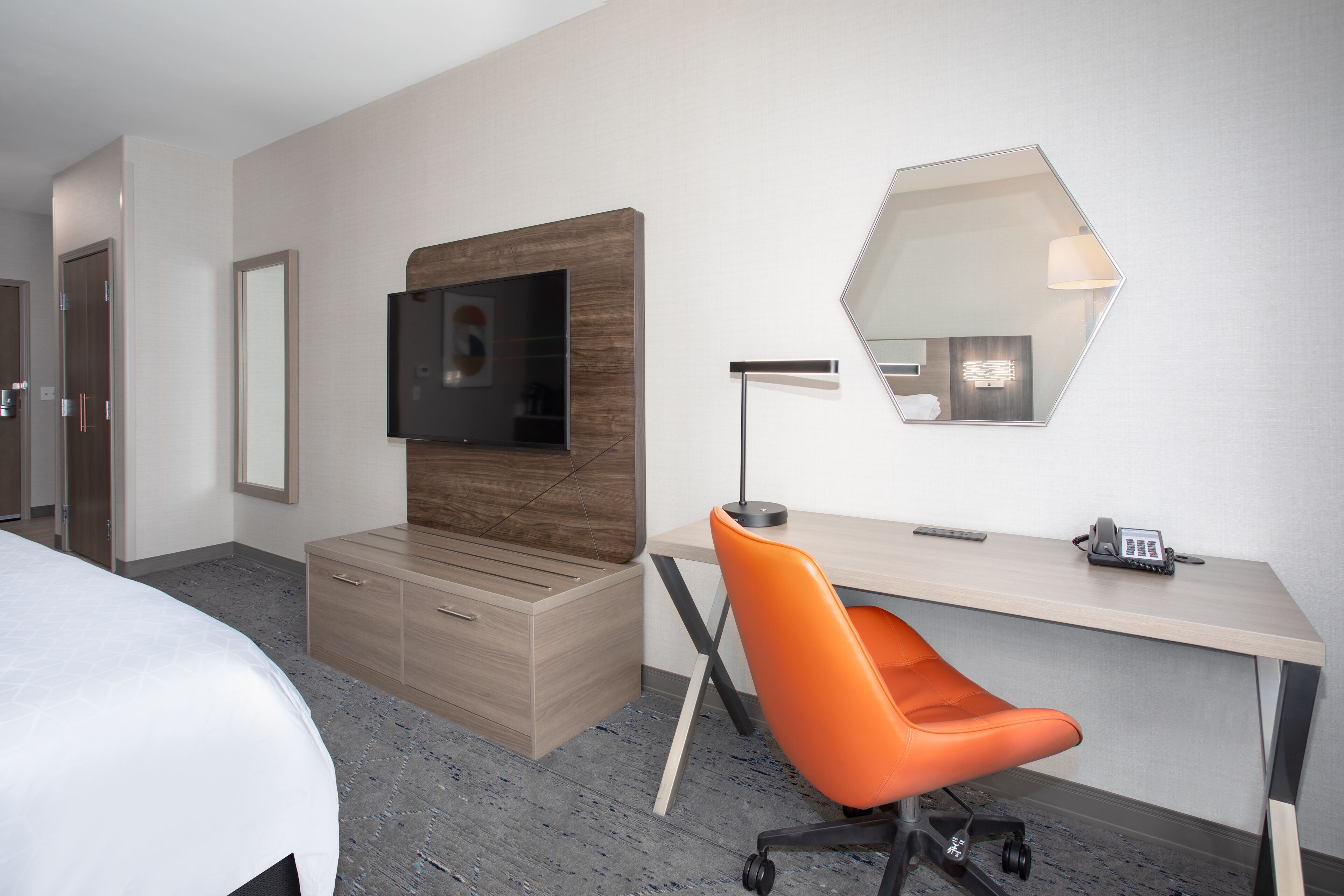 Habitación de hotel con cama, un escritorio con silla, televisor montado en la pared, un espejo y un teléfono.