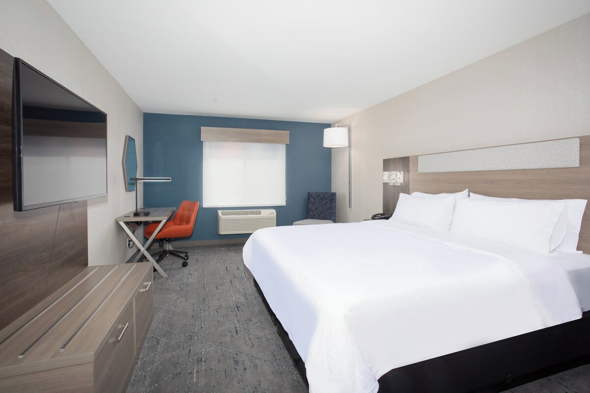 A hotel room with a bed, a desk, an orange chair, a flat-screen TV, and a window with blinds.