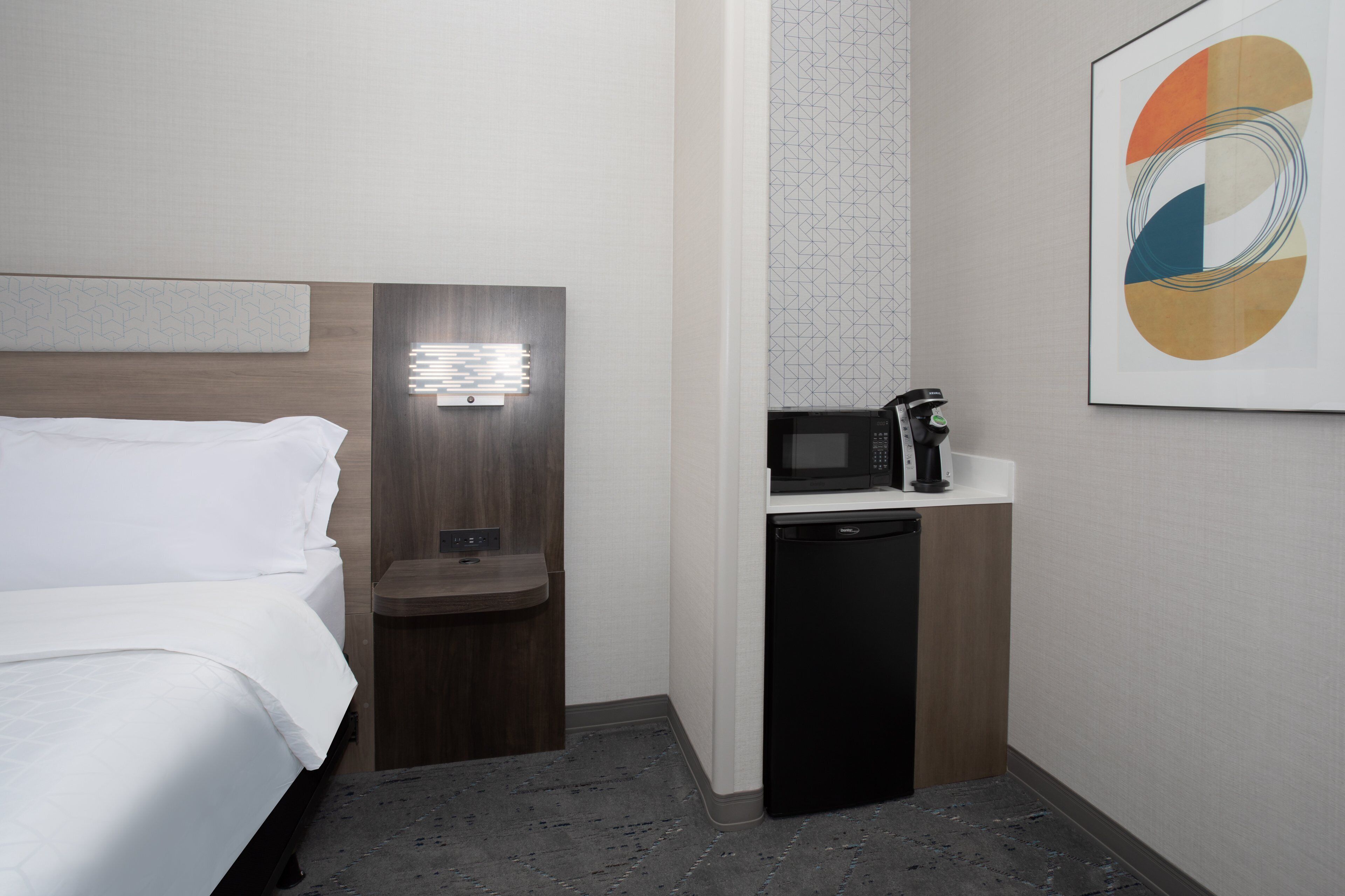 A hotel room with a bed, bedside table, microwave, and a small refrigerator.