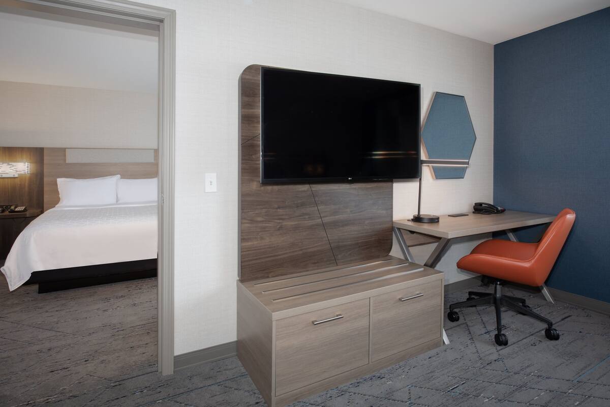 A hotel room with a bed, a desk with a chair, a flat-screen TV, and a wall-mounted lamp.