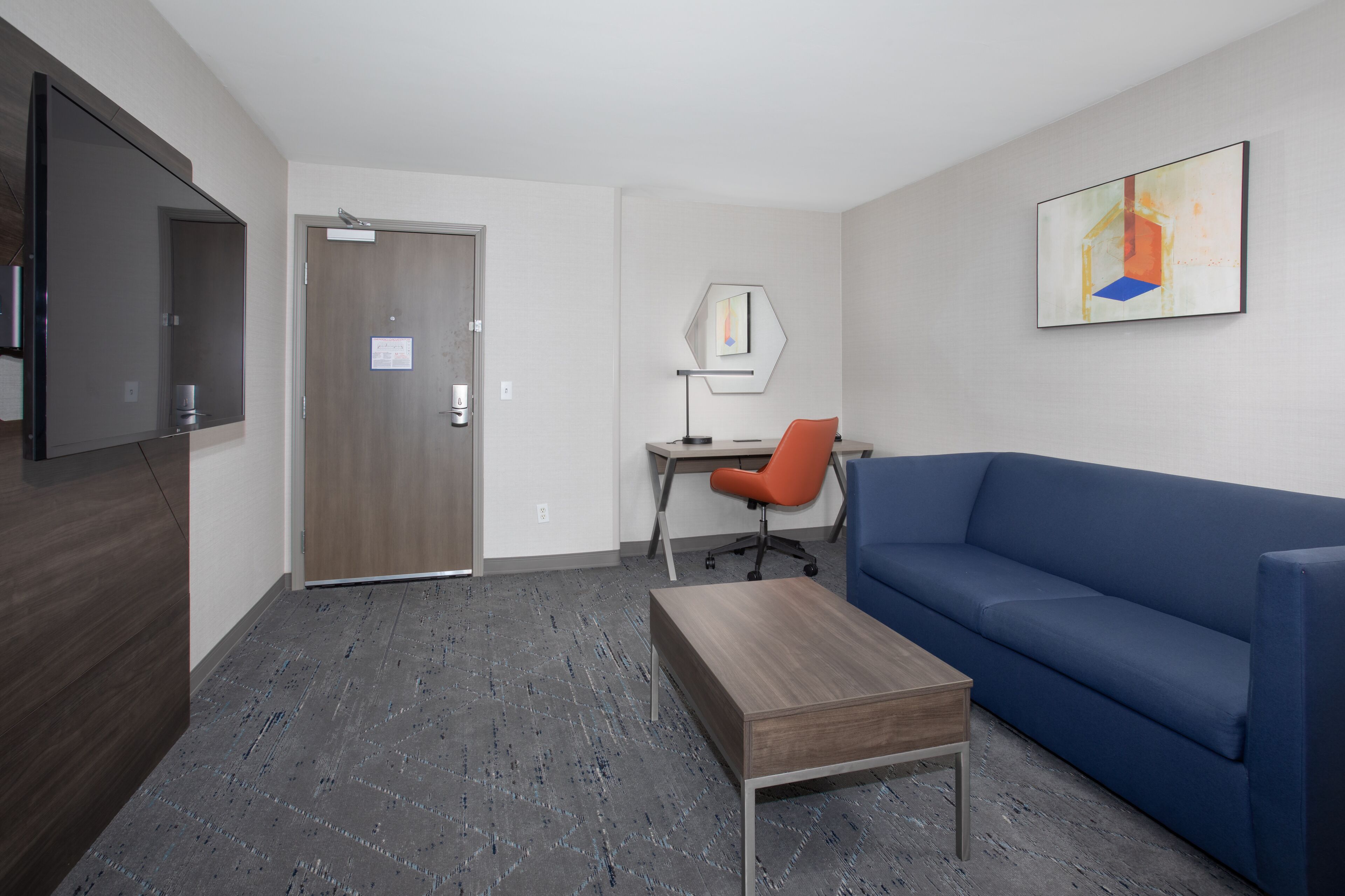 Una habitació d'hotel moderna amb escriptori de fusta, sofà blau, televisió de pantalla plana i un quadre a la paret.