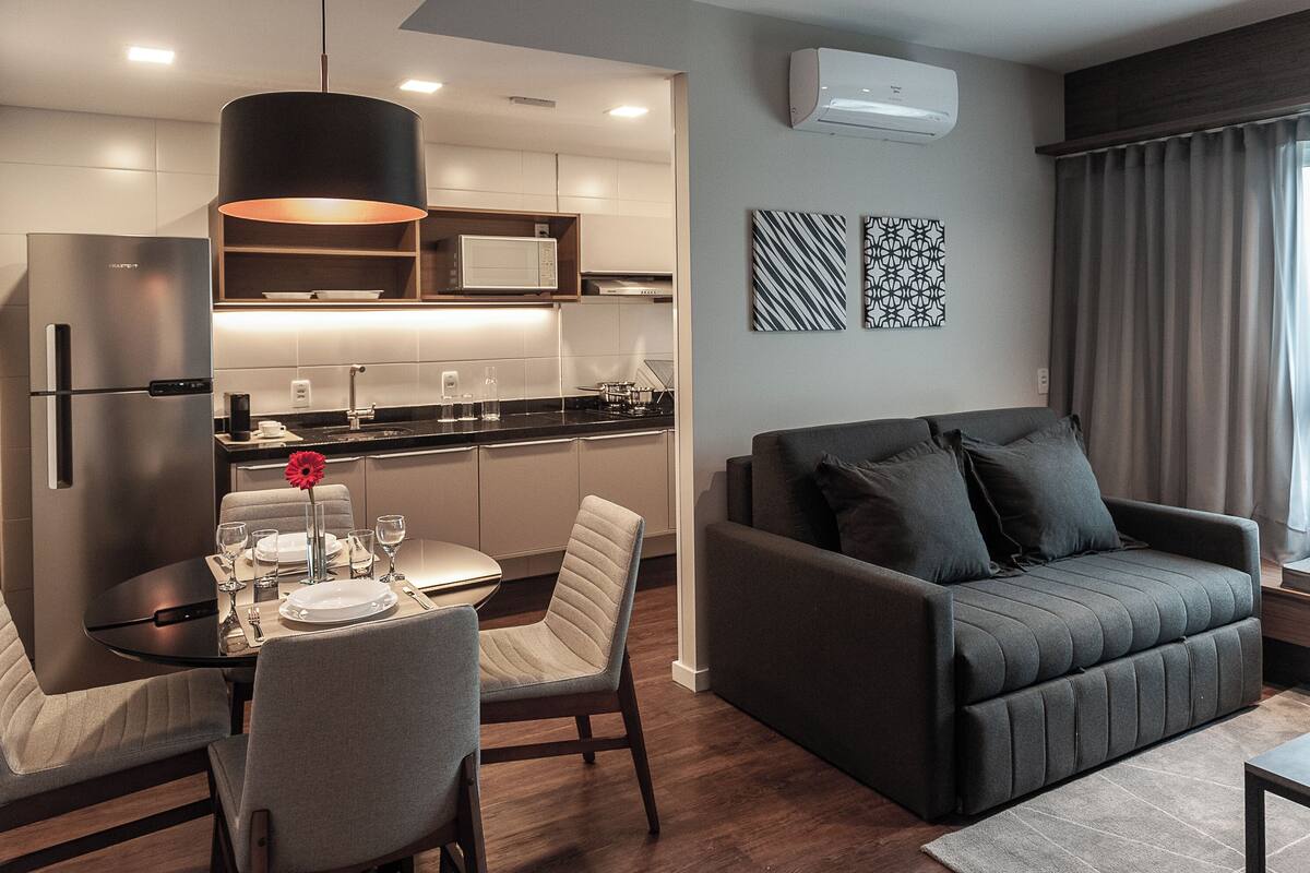 Una sala moderna con un sofá gris oscuro, una mesa de comedor con platos y vasos, una zona de cocina con microondas y un ventanal con cortinas.