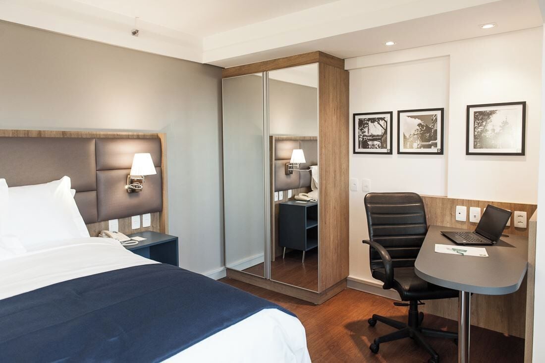Een hotelkamer met een bed, nachtkastjes, een bureau met stoel, een laptop en ingekaderde foto's aan de muur.