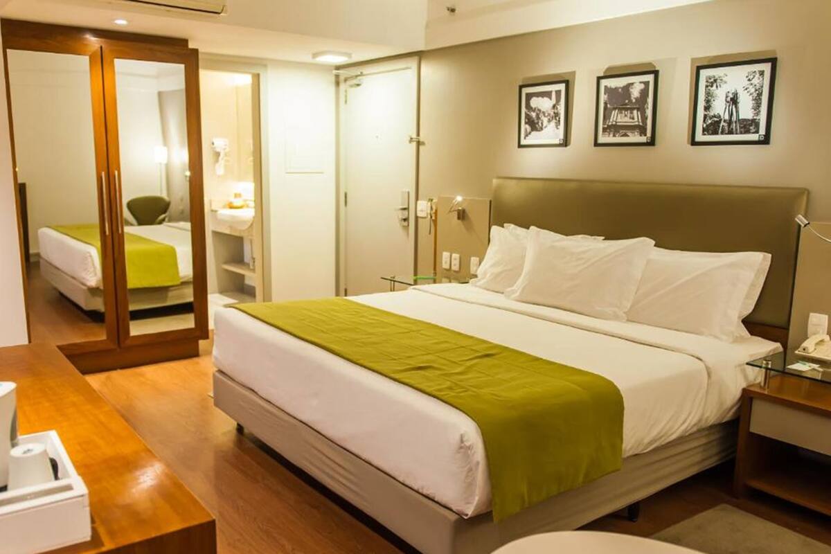 Una habitación de hotel con cama, escritorio, silla, espejo, armario y mesita de noche.