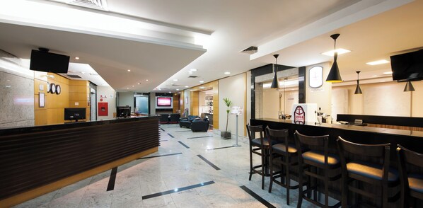 Um hall moderno de hotel com balcão de recepção, área de bar, assentos e uma tela de televisão.