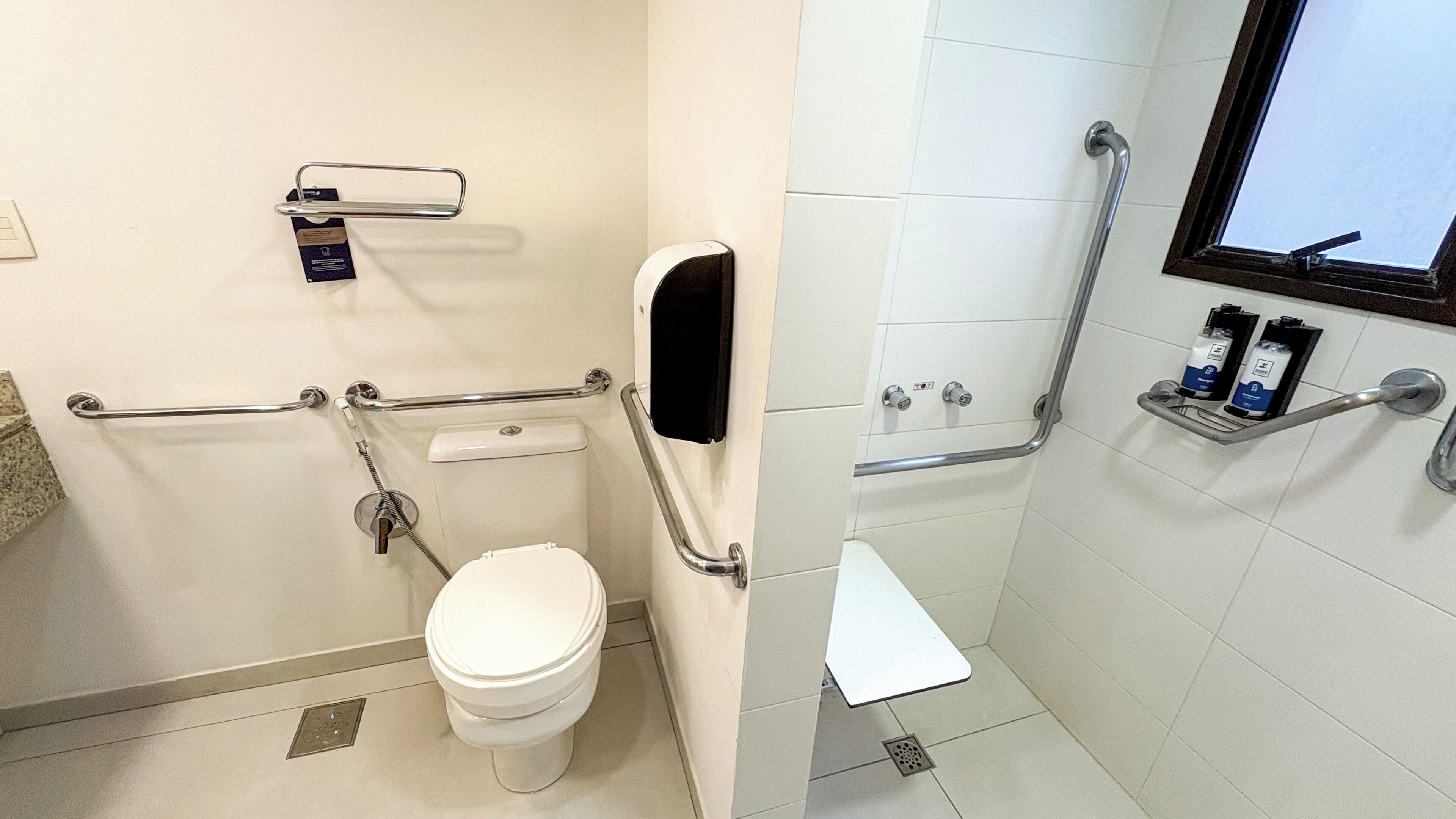 Un baño con inodoro, barras de apoyo, una ventana y un estante con artículos de aseo.