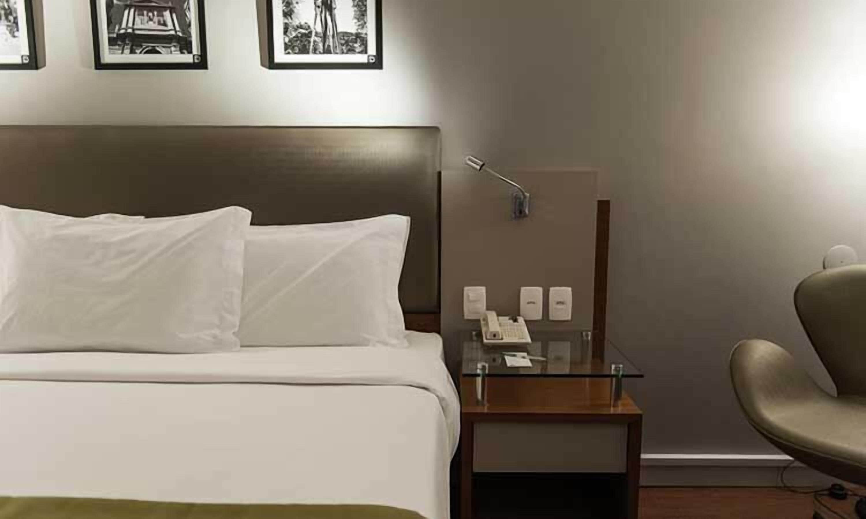A hotel room with a bed, a nightstand with a lamp, a chair, and a wall-mounted telephone.