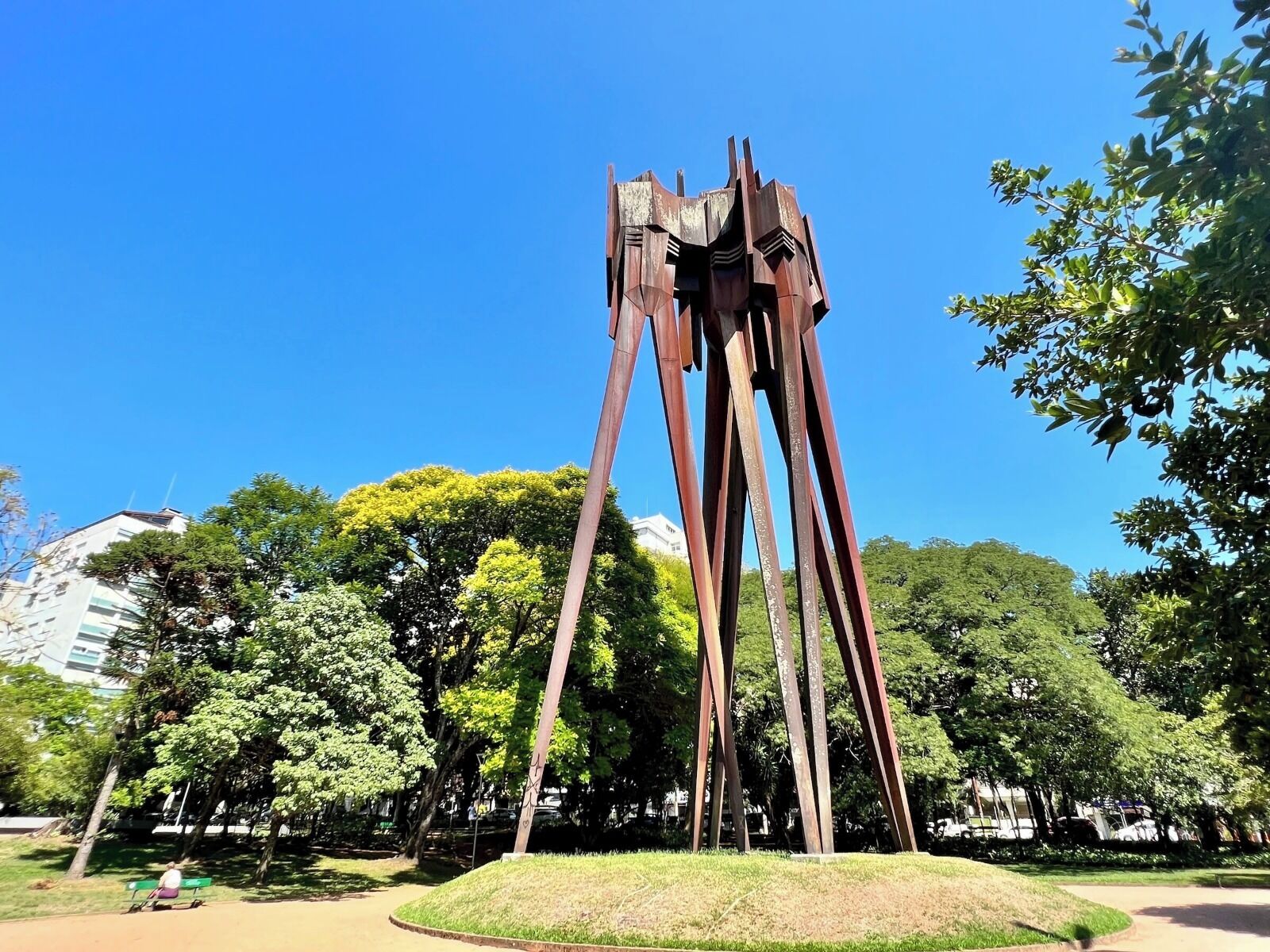 A tall, abstract metal sculpture in a park with trees and a clear blue sky.