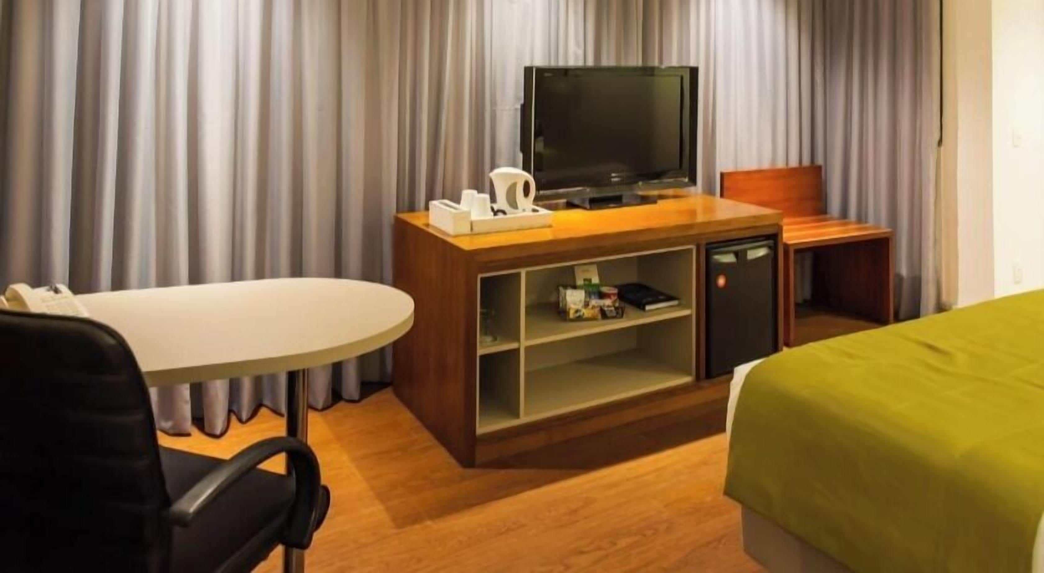 Quarto de hotel com escrivaninha de madeira, televisão, minibar e cama com cobertura verde.