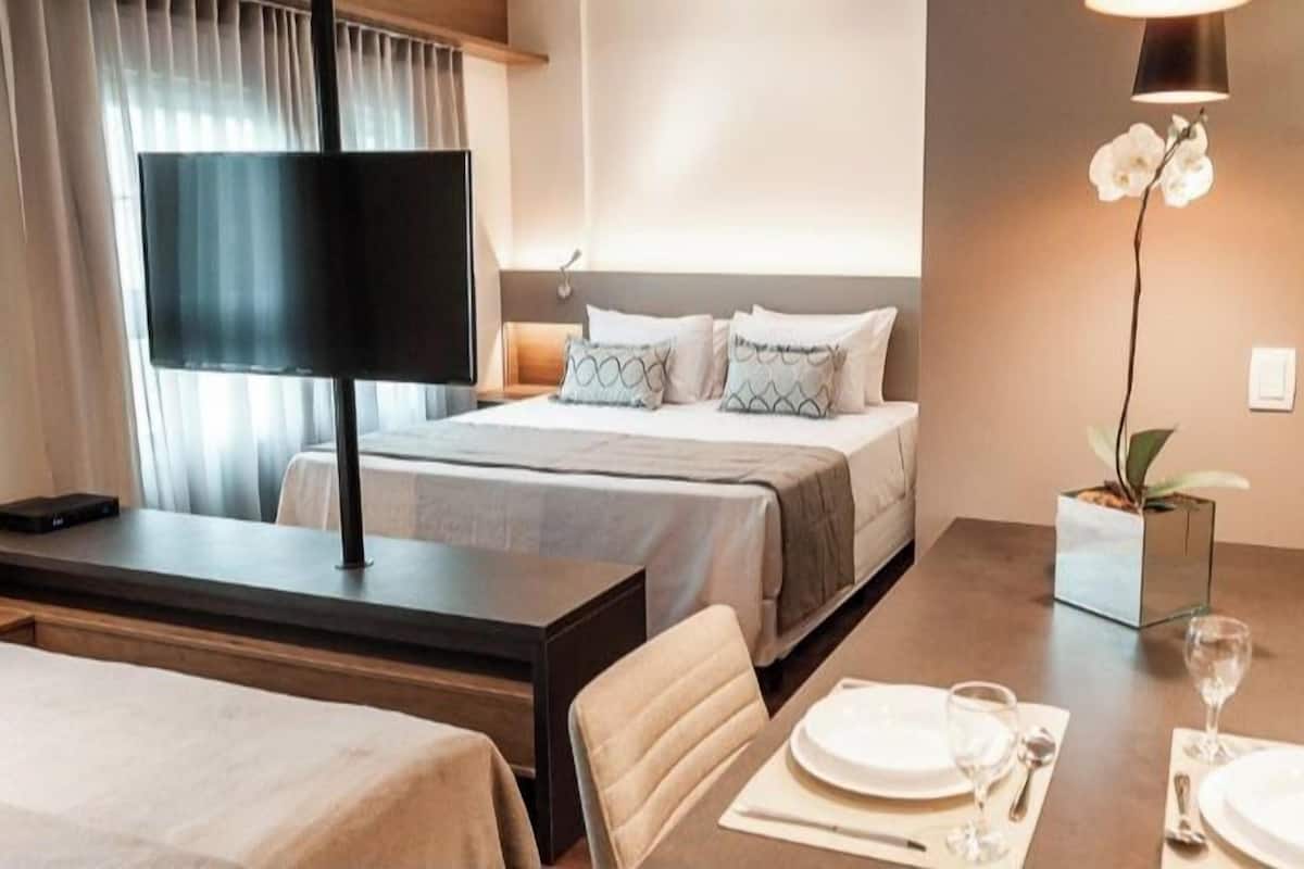 Una habitación de hotel con cama, mesita de noche, lámpara y un rincón comedor con una mesa dispuesta para dos personas.