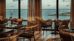 A restaurant with wicker furniture, a checkered floor, and a view of the sea through large windows. - AX ODYCY Hotel (St. Paul's Bay)