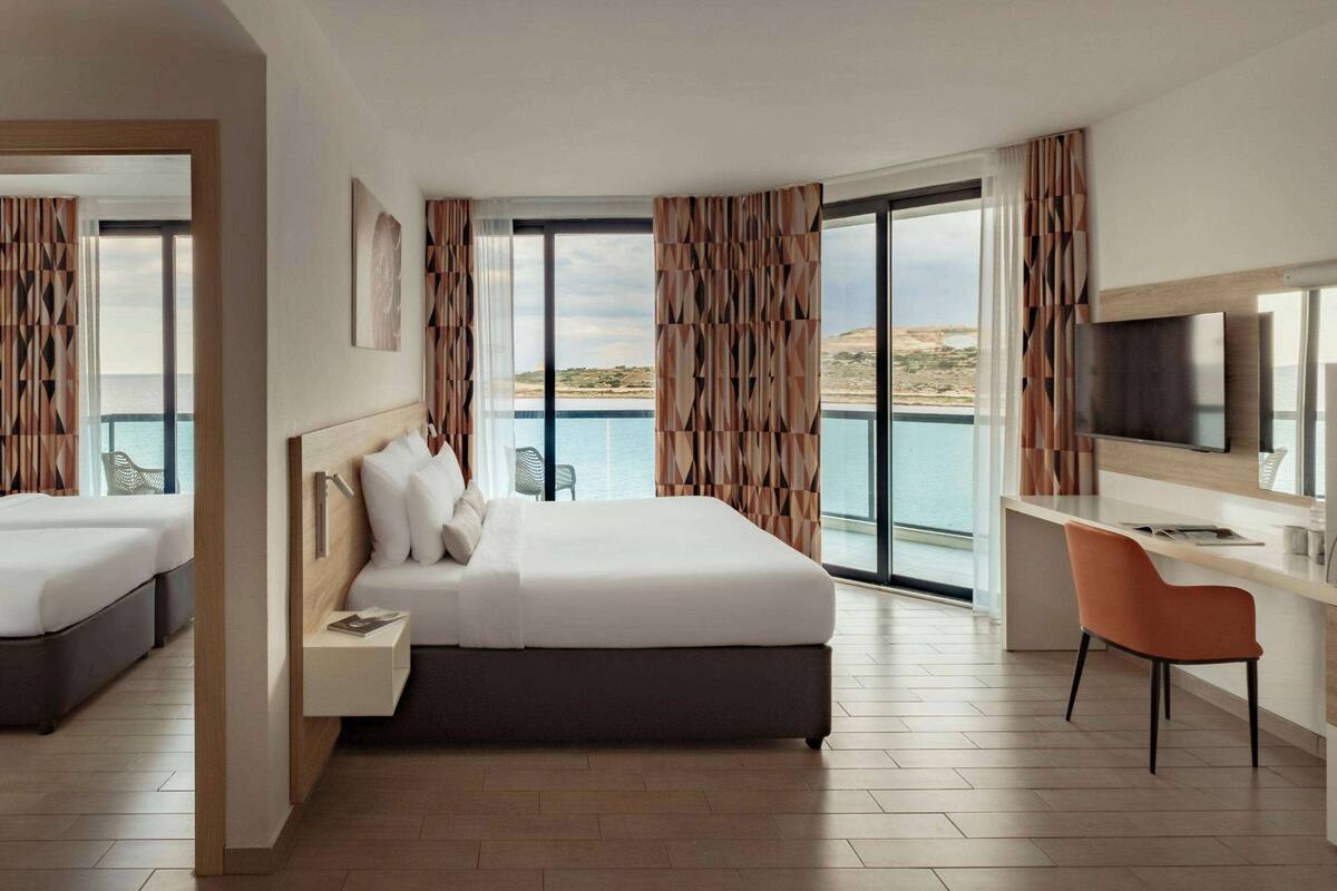 A hotel room with a bed, a desk, a chair, and a TV. The room has a large window with a view of the sea.