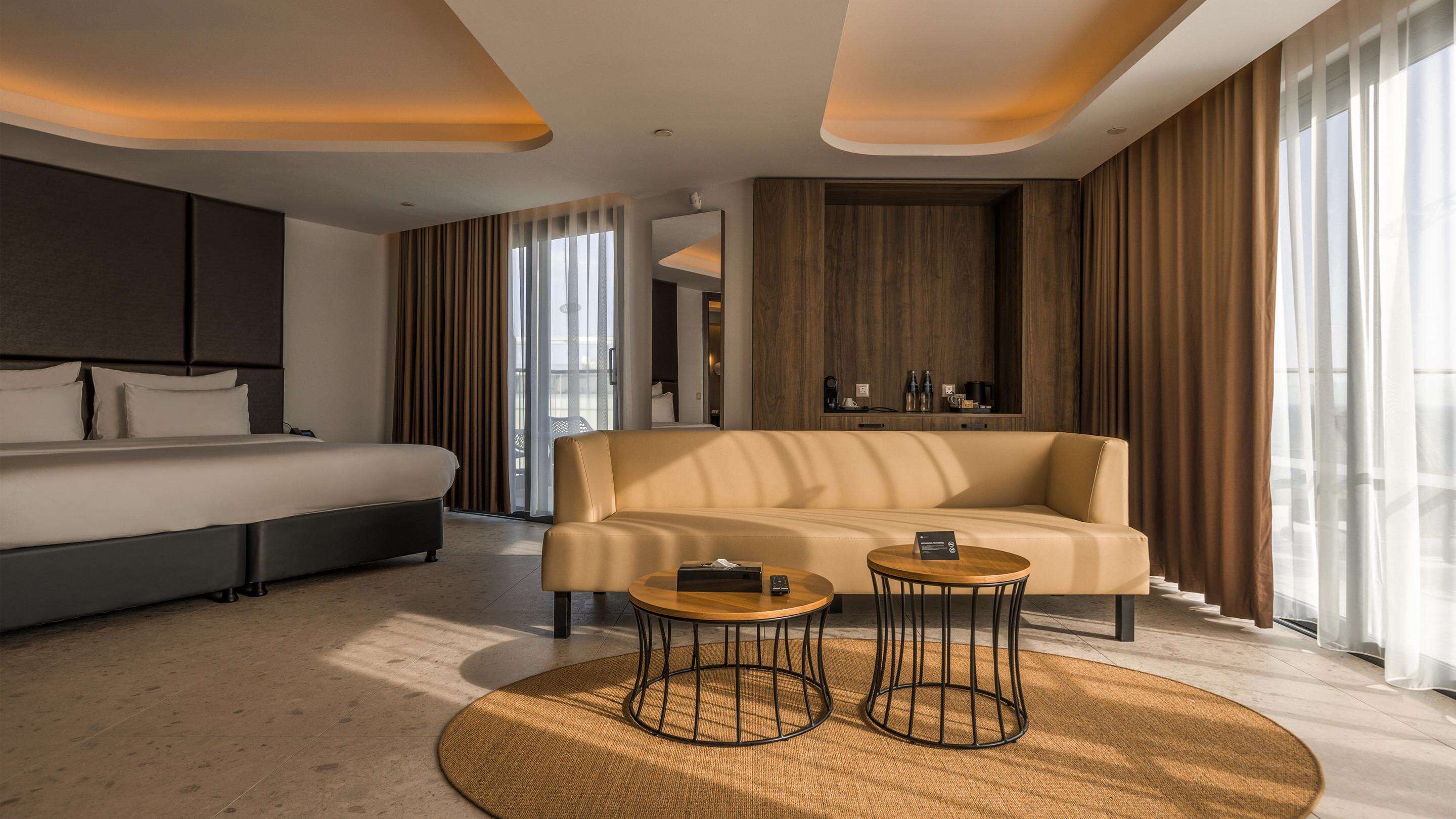 A modern hotel room with a large bed, a sofa, two small tables, and a large window with curtains.