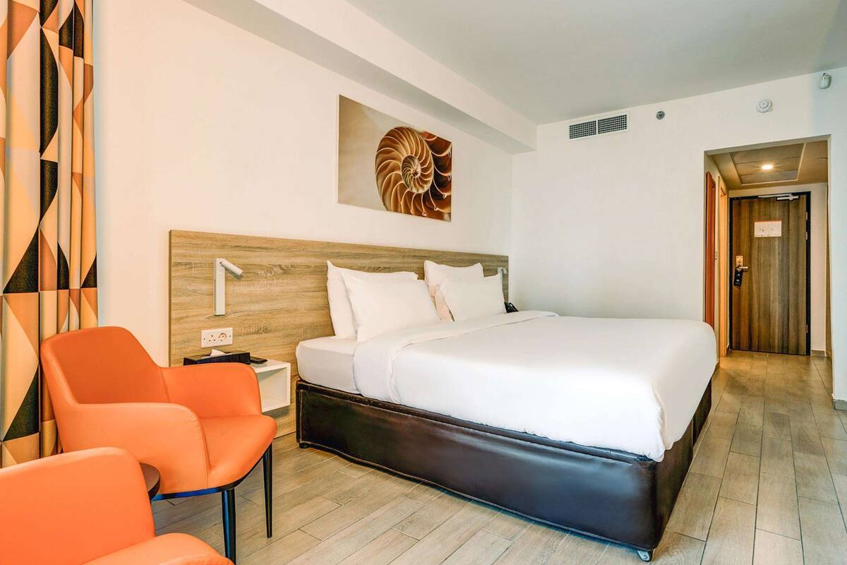 A modern hotel room with a large bed, orange chairs, a wooden headboard, and a wall-mounted artwork.