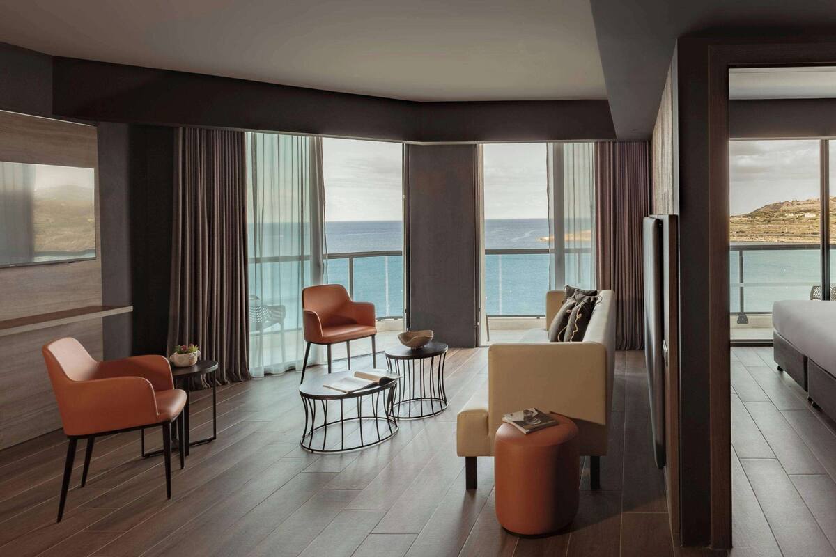 A modern hotel room with a large balcony, a bed, two armchairs, a coffee table, and a view of the sea.
