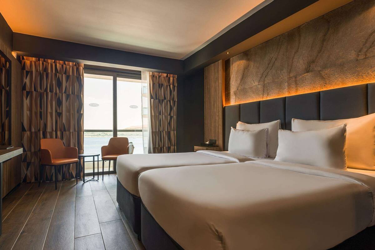 A hotel room with two beds, a TV, a desk, and a view of the sea.