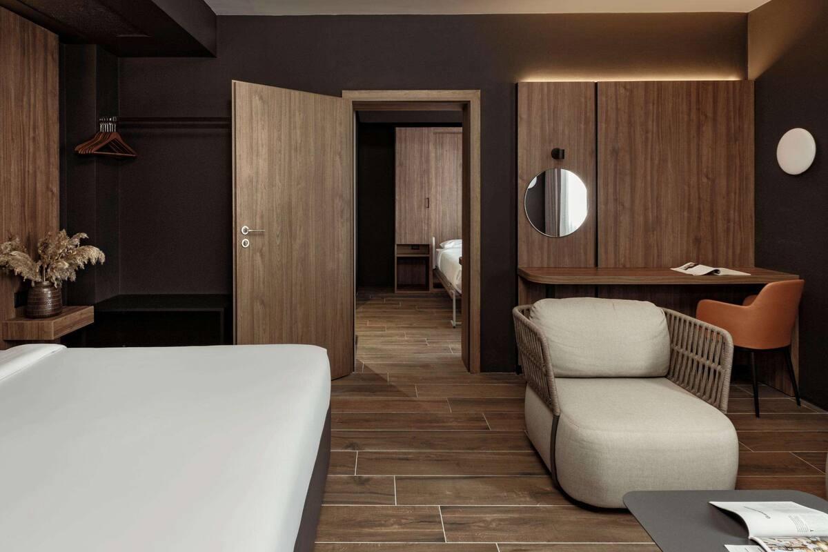 A modern hotel room with a large bed, a seating area with a chair, a small desk, and a mirror.