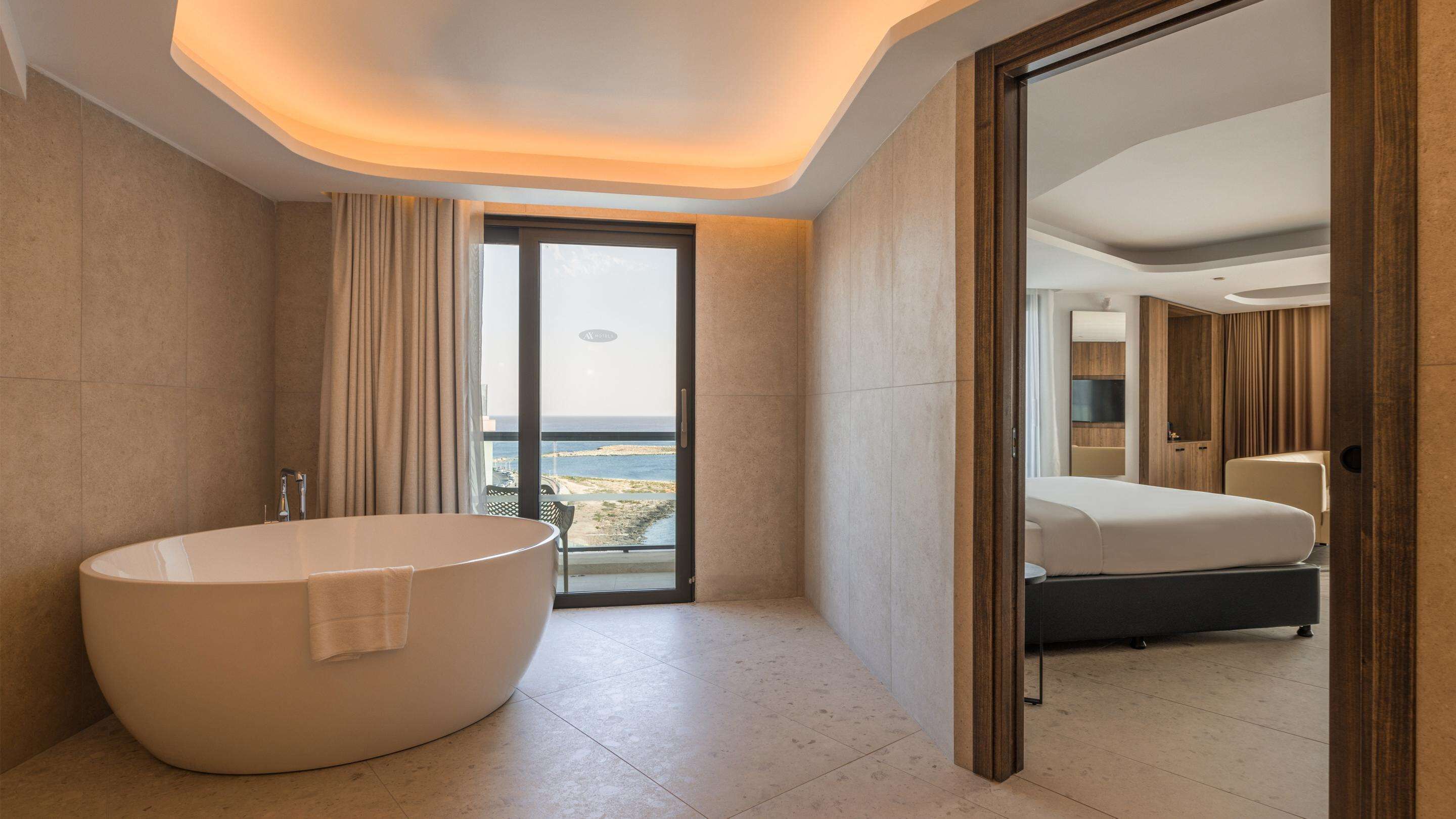 Un baño moderno con una bañera independiente, un espejo grande y vista al mar a través de una puerta corredera.