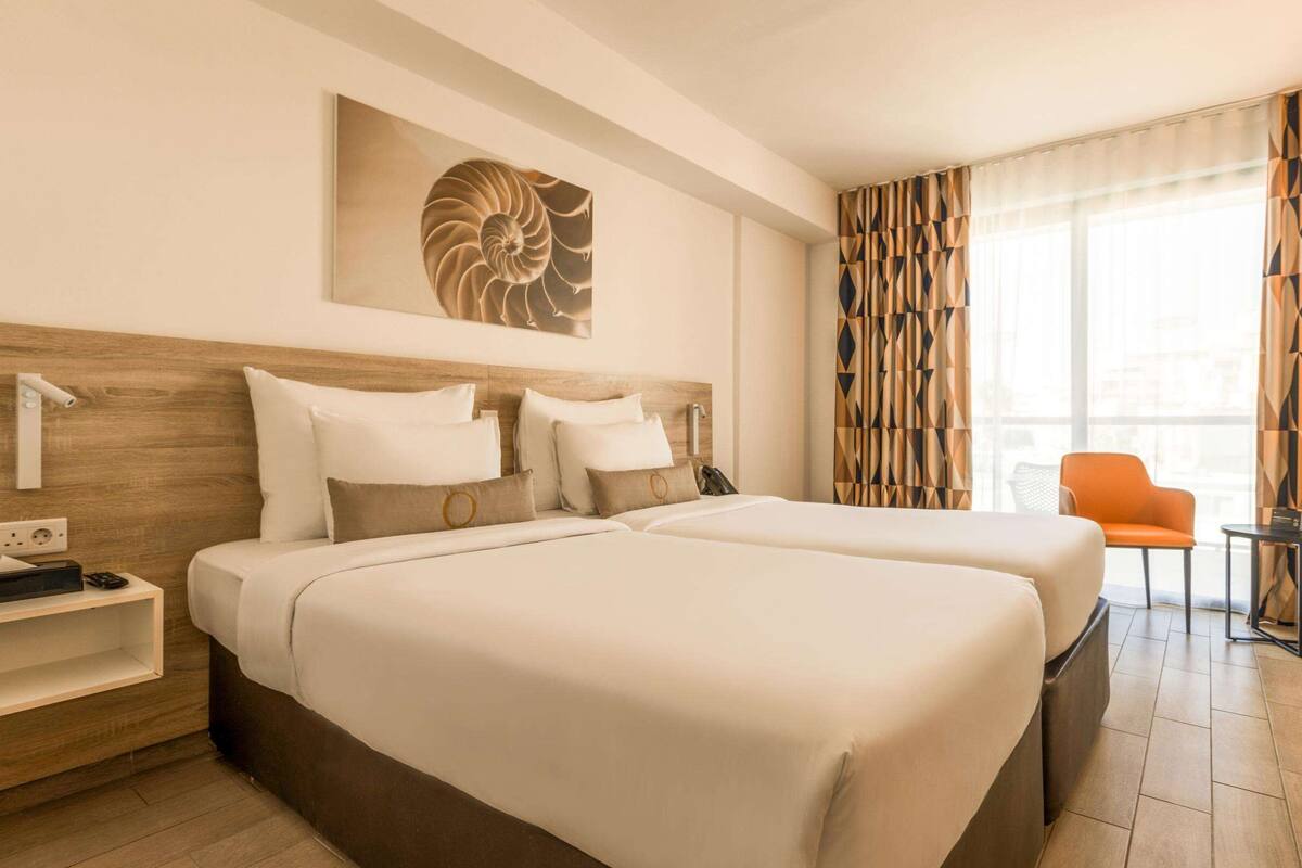 A hotel room with two beds, a wooden headboard, a wall-mounted artwork, a bedside table with a phone and a radio, and a window with patterned curtains.