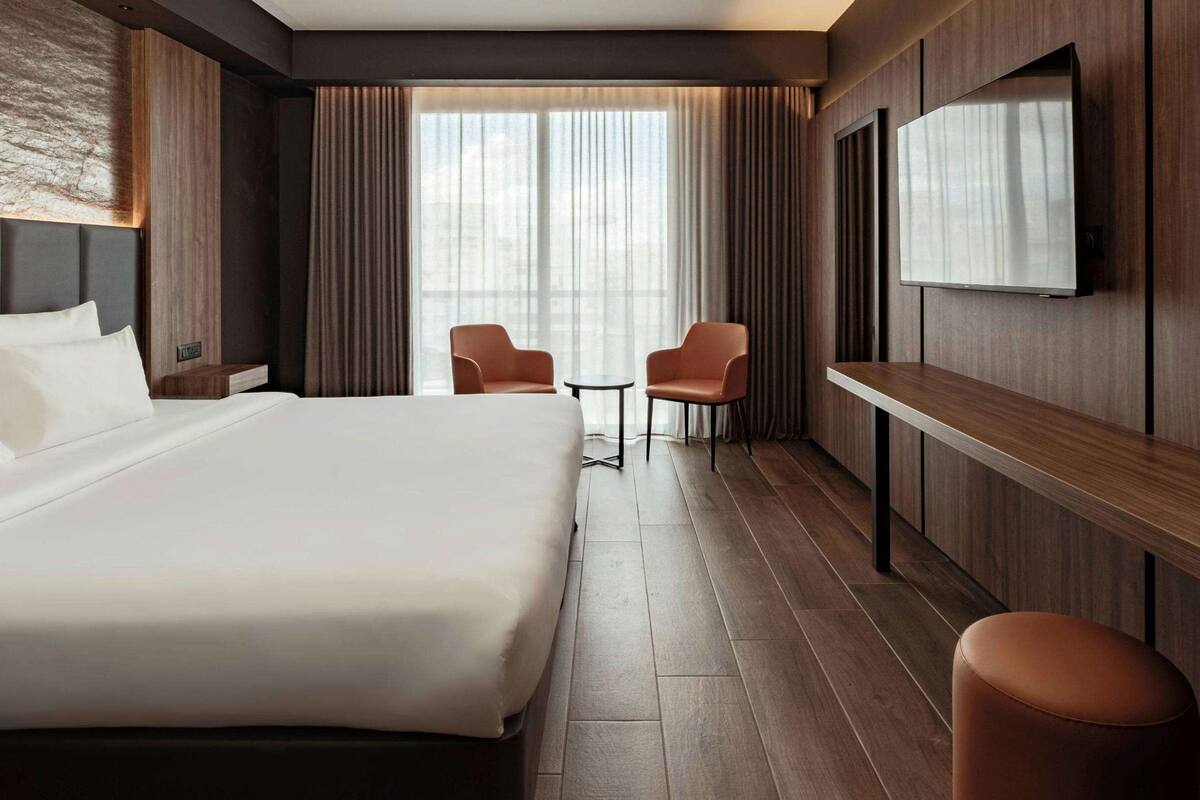 A modern hotel room with a large bed, two chairs, a small table, a TV, and a large window with curtains.